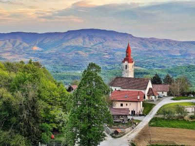 A white church with a red-tiled roof and steeple stands in a green, rural landscape with rolling hills and mountains in the background.