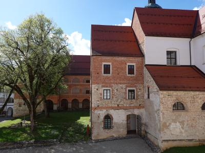 A courtyard view of a historic monastery complex featuring a brick and stone building with a red tiled roof, an arched cloister walkway, and a large tree on a grassy lawn under a blue sky.