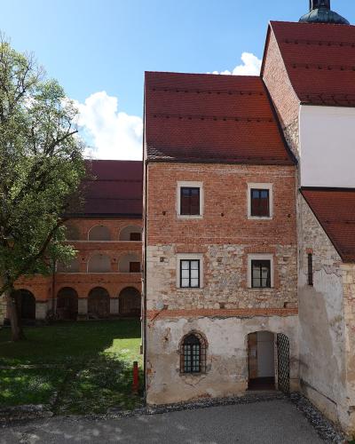 A courtyard view of a historic monastery complex featuring a brick and stone building with a red tiled roof, an arched cloister walkway, and a large tree on a grassy lawn under a blue sky.