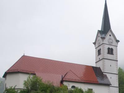 A white church with a red-tiled roof and a tall, slender steeple featuring clock faces, set against an overcast sky with green trees in the foreground.