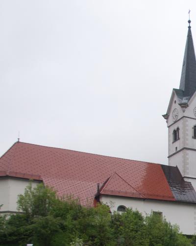 A white church with a red-tiled roof and a tall, slender steeple featuring clock faces, set against an overcast sky with green trees in the foreground.