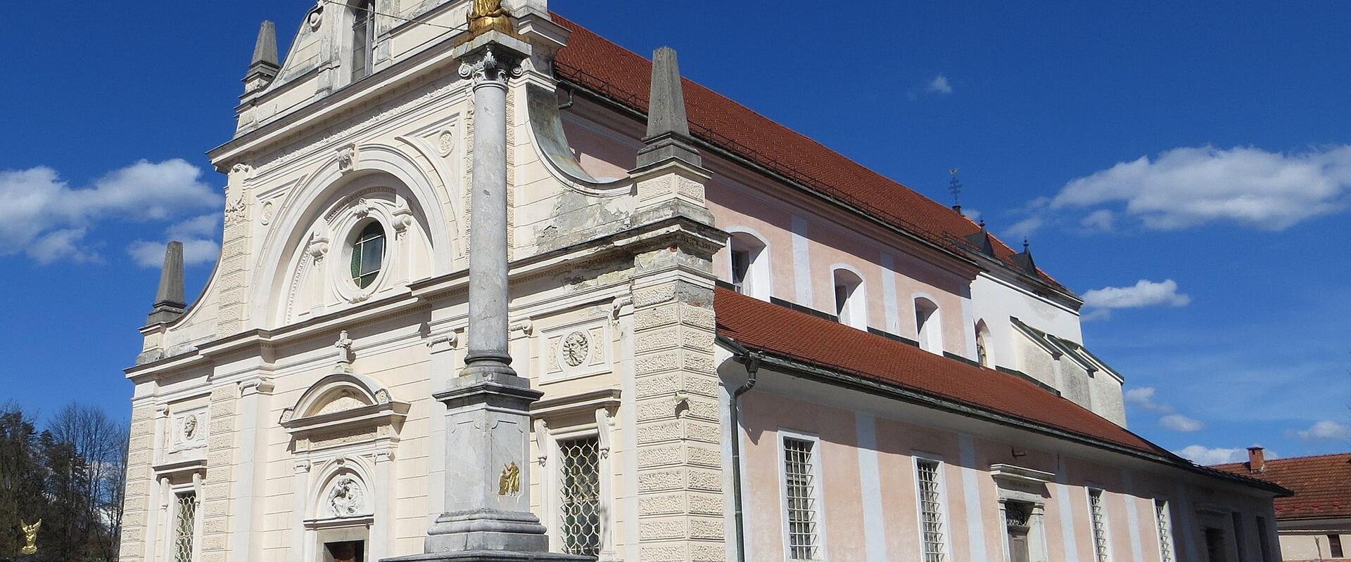 A white Baroque-style parish church with a prominent golden statue of a saint on a column in the foreground and a separate stone bell tower to the right, set in a paved square under a clear blue sky.