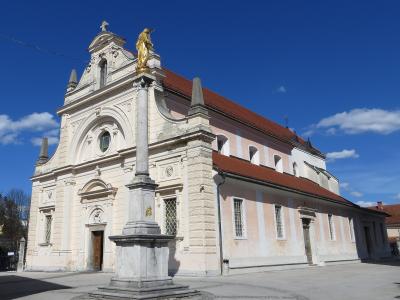 A white Baroque-style parish church with a prominent golden statue of a saint on a column in the foreground and a separate stone bell tower to the right, set in a paved square under a clear blue sky.