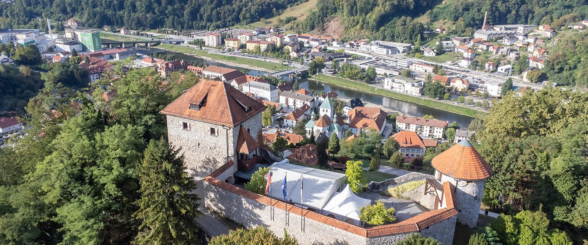 An aerial view of Laško Castle in the foreground, overlooking the town of Laško, the Savinja River, and the historic Laško Brewery complex in the background.