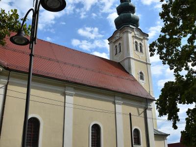 A low-angle photograph of a cream-colored Baroque-style church with a red-tiled roof and a distinctive multi-tiered onion dome bell tower, framed by trees and a street lamp.