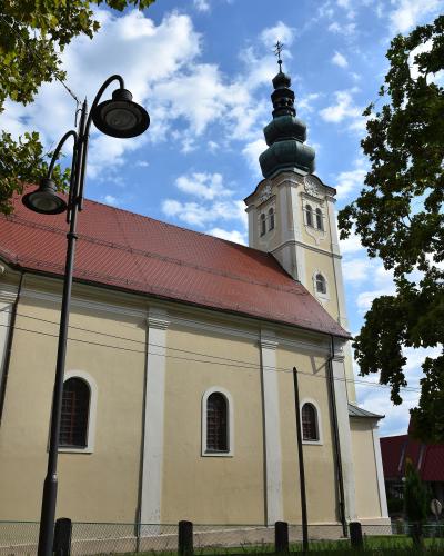 A low-angle photograph of a cream-colored Baroque-style church with a red-tiled roof and a distinctive multi-tiered onion dome bell tower, framed by trees and a street lamp.