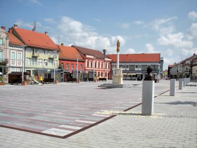 A wide, paved town square featuring a central plague column with a golden statue, a bronze bust on a pedestal in the foreground, and colorful historic buildings lining the perimeter under a blue sky.