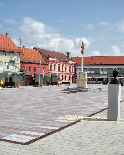 A wide, paved town square featuring a central plague column with a golden statue, a bronze bust on a pedestal in the foreground, and colorful historic buildings lining the perimeter under a blue sky.