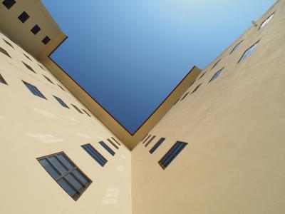 A low-angle architectural photograph looking up into a courtyard or corner formed by two tall, pale yellow stucco walls with rectangular windows under a clear blue sky.