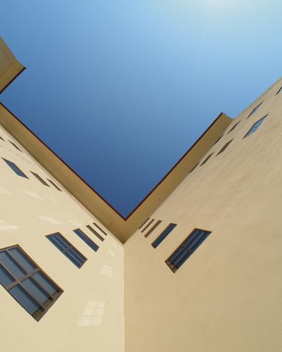 A low-angle architectural photograph looking up into a courtyard or corner formed by two tall, pale yellow stucco walls with rectangular windows under a clear blue sky.