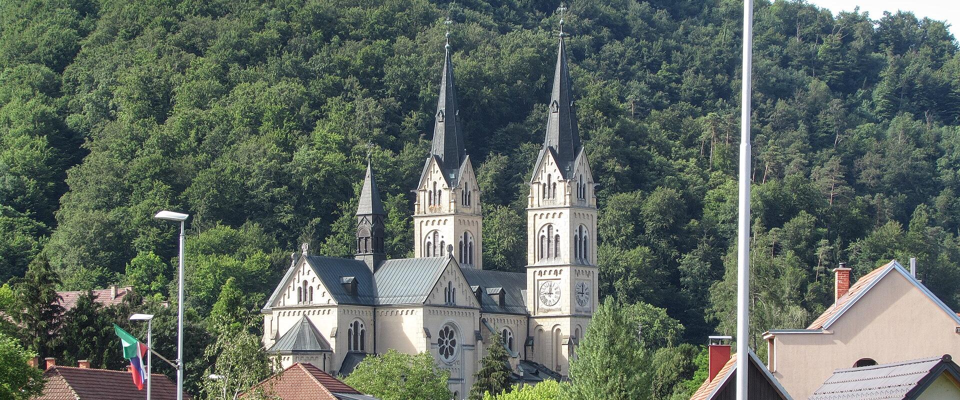 A large Neo-Romanesque church with two prominent spires and a smaller central tower, set against a densely forested hillside, with residential houses and a Slovenian flag in the foreground.