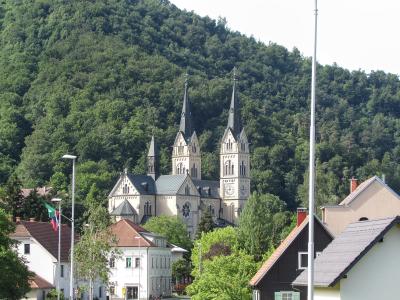 A large Neo-Romanesque church with two prominent spires and a smaller central tower, set against a densely forested hillside, with residential houses and a Slovenian flag in the foreground.