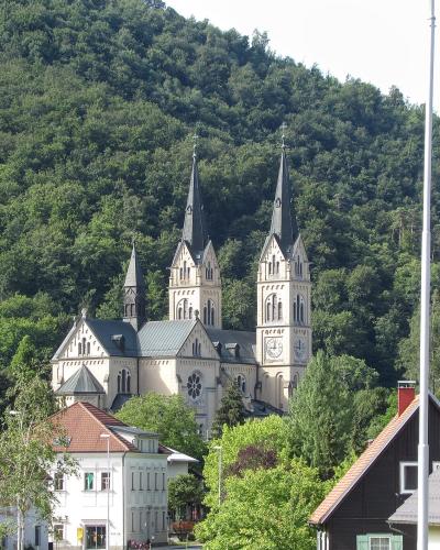 A large Neo-Romanesque church with two prominent spires and a smaller central tower, set against a densely forested hillside, with residential houses and a Slovenian flag in the foreground.