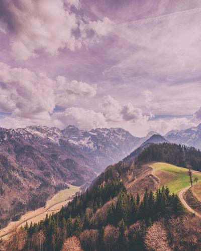 A high-angle landscape photograph showing a winding dirt road leading up a forested hillside to a cluster of buildings, with a deep valley and snow-dusted mountain range in the background under a cloudy sky.