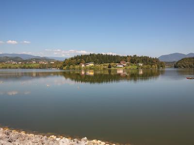 A wide landscape shot of a calm lake with a rocky embankment in the foreground, a tree-covered peninsula in the middle distance, and rolling hills under a blue sky.