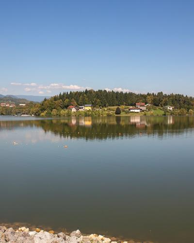 A wide landscape shot of a calm lake with a rocky embankment in the foreground, a tree-covered peninsula in the middle distance, and rolling hills under a blue sky.