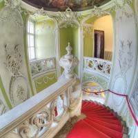 The image shows an ornate, curved interior staircase with a red carpet runner, white Rococo-style plasterwork, pale green walls, and a painted ceiling fresco, viewed from above looking down the stairs.
