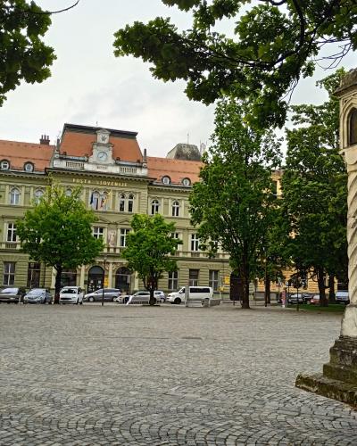 A cobblestone square featuring a prominent twisted stone column (the Plague Column) in the foreground, with a large yellow building labeled "POSTA SLOVENIJE" (Slovenian Post) in the background, framed by green trees.