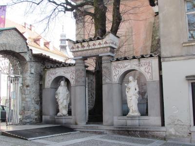 A stone architectural structure with two arched niches containing white statues, featuring decorative red and grey patterns on the arches and a tiled roof, situated outdoors next to a stone wall and tree.