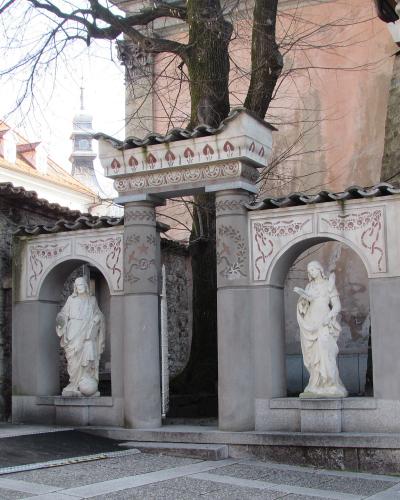 A stone architectural structure with two arched niches containing white statues, featuring decorative red and grey patterns on the arches and a tiled roof, situated outdoors next to a stone wall and tree.