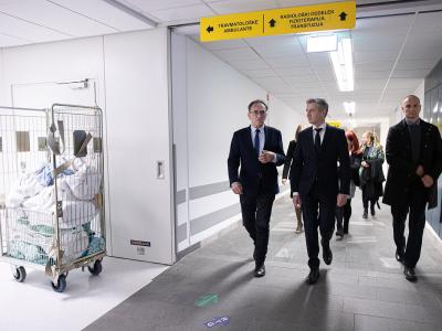 The image shows a group of men in suits walking down a hospital corridor, with a yellow overhead sign directing to "TRAVMATOLOŠKE AMBULANTE" (Traumatology Ambulances) and "RADIOLOŠKI ODDELEK" (Radiology Department), alongside a laundry cart on the le