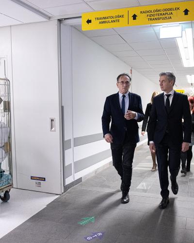 The image shows a group of men in suits walking down a hospital corridor, with a yellow overhead sign directing to "TRAVMATOLOŠKE AMBULANTE" (Traumatology Ambulances) and "RADIOLOŠKI ODDELEK" (Radiology Department), alongside a laundry cart on the le