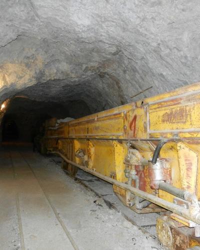 A view inside a rough-hewn underground tunnel or mine shaft featuring a large piece of yellow industrial mining machinery on rails.