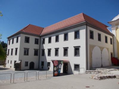 A white, multi-story building with a red-tiled roof, featuring a modern entrance canopy with colorful circular graphics and a small stone ruin in the foreground.