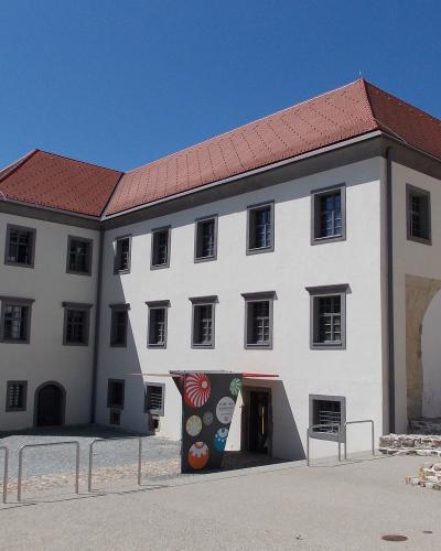 A white, multi-story building with a red-tiled roof, featuring a modern entrance canopy with colorful circular graphics and a small stone ruin in the foreground.