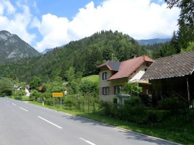 A paved road winding through a green valley with houses and forested mountains in the background, featuring a yellow rectangular road sign on the left side of the road.