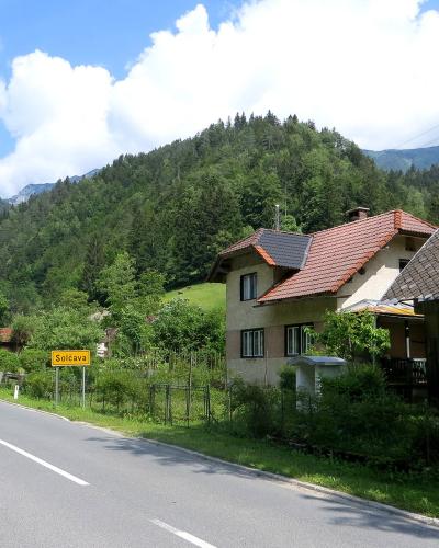 A paved road winding through a green valley with houses and forested mountains in the background, featuring a yellow rectangular road sign on the left side of the road.