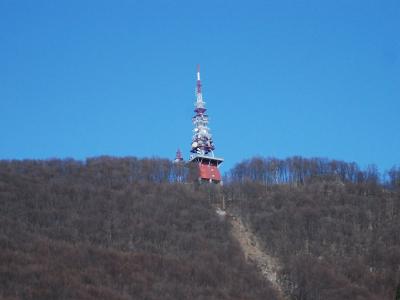 A tall telecommunications tower with red and white markings stands on the ridge of a forested hill under a clear blue sky.