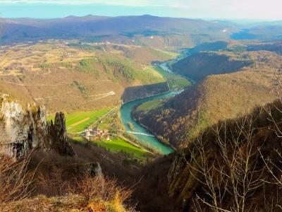 A high-angle landscape photograph showing a winding turquoise river flowing through a deep valley with green fields and forests, viewed from a rocky cliff edge with bare trees in the foreground.