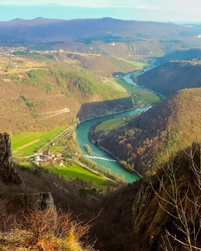 A high-angle landscape photograph showing a winding turquoise river flowing through a deep valley with green fields and forests, viewed from a rocky cliff edge with bare trees in the foreground.