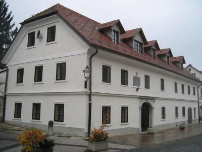 A two-story white building with a red-tiled roof, dormer windows, and a plaque on the facade, situated on a paved street with planters and a lamppost.