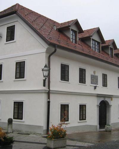 A two-story white building with a red-tiled roof, dormer windows, and a plaque on the facade, situated on a paved street with planters and a lamppost.
