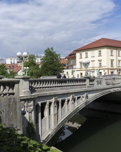 A stone arch bridge with prominent green dragon statues at its corners, spanning a river in an urban setting with European-style buildings in the background; visible inscriptions include "1848" and "1895" on the bridge’s parapet.