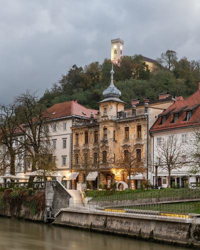 A riverside view of historic buildings along the Ljubljanica River with Ljubljana Castle visible on the hilltop in the background.