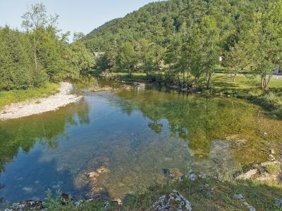 A clear, shallow river flows through a green, wooded valley with a forested hill in the background and a small settlement visible on the far bank.