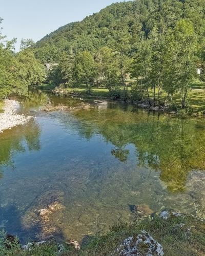 A clear, shallow river flows through a green, wooded valley with a forested hill in the background and a small settlement visible on the far bank.