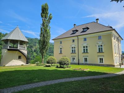 A yellow Baroque-style manor house with white window frames and a dark tiled roof, situated on a green lawn next to a distinctive octagonal pavilion with an external spiral staircase, set against a backdrop of forested hills under a blue sky.