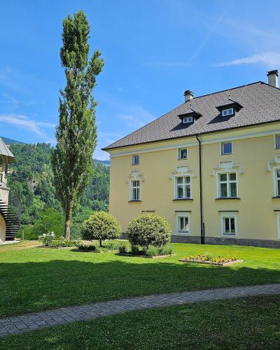 A yellow Baroque-style manor house with white window frames and a dark tiled roof, situated on a green lawn next to a distinctive octagonal pavilion with an external spiral staircase, set against a backdrop of forested hills under a blue sky.