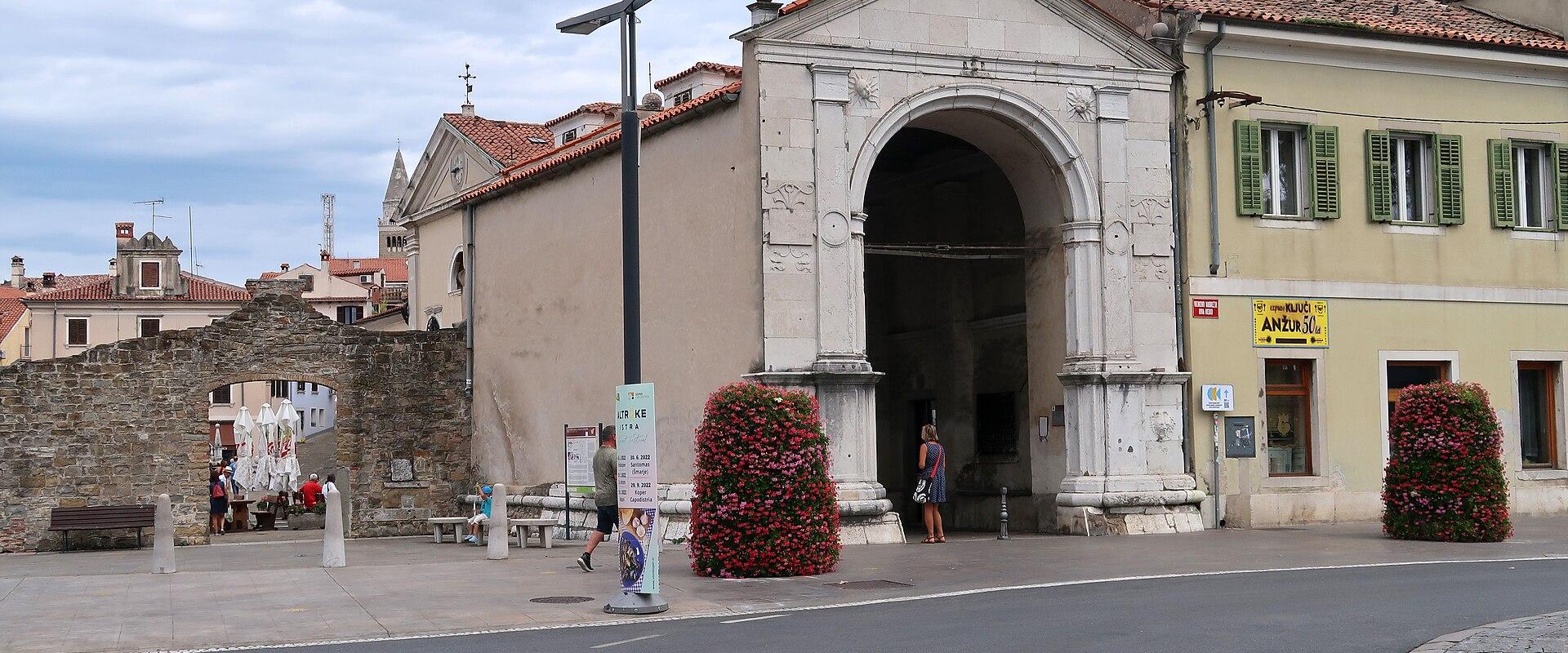 A large Renaissance-style stone archway with a triangular pediment, flanked by a yellow building with green shutters and a smaller stone ruin, situated in a paved town square.