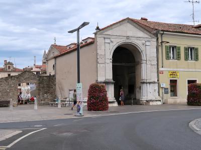 A large Renaissance-style stone archway with a triangular pediment, flanked by a yellow building with green shutters and a smaller stone ruin, situated in a paved town square.