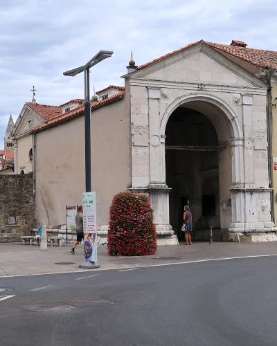 A large Renaissance-style stone archway with a triangular pediment, flanked by a yellow building with green shutters and a smaller stone ruin, situated in a paved town square.
