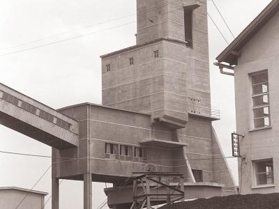 A black and white photograph showing a large, multi-story concrete headframe (winding tower) of a mine, with a covered conveyor bridge extending from its side and a railway freight car filled with coal in the foreground.