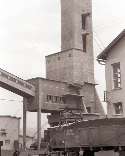A black and white photograph showing a large, multi-story concrete headframe (winding tower) of a mine, with a covered conveyor bridge extending from its side and a railway freight car filled with coal in the foreground.