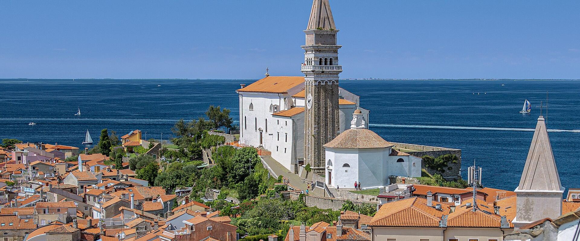 A high-angle view of a coastal town with terracotta rooftops, dominated by a large white church with a tall, octagonal stone bell tower topped by a statue, overlooking the blue sea.