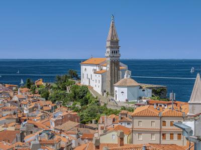 A high-angle view of a coastal town with terracotta rooftops, dominated by a large white church with a tall, octagonal stone bell tower topped by a statue, overlooking the blue sea.