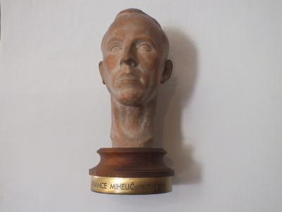 A terracotta bust of a man mounted on a wooden base with a brass plaque reading "FRANCE MIHELIČ 1907 - 1998" against a plain white wall.